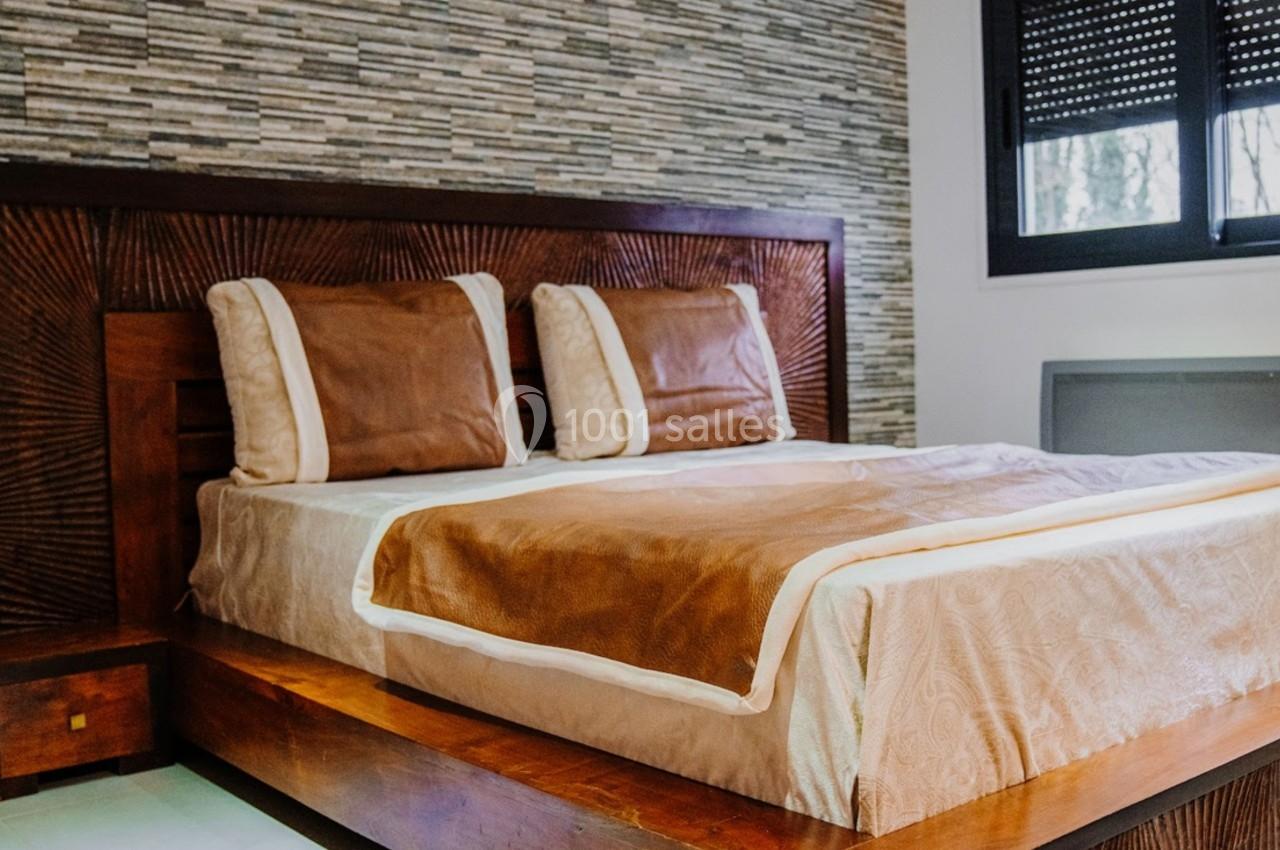 Lit double avec cadre en bois foncé, draps beige et marron, devant un mur en pierre et une fenêtre avec volet roulant.