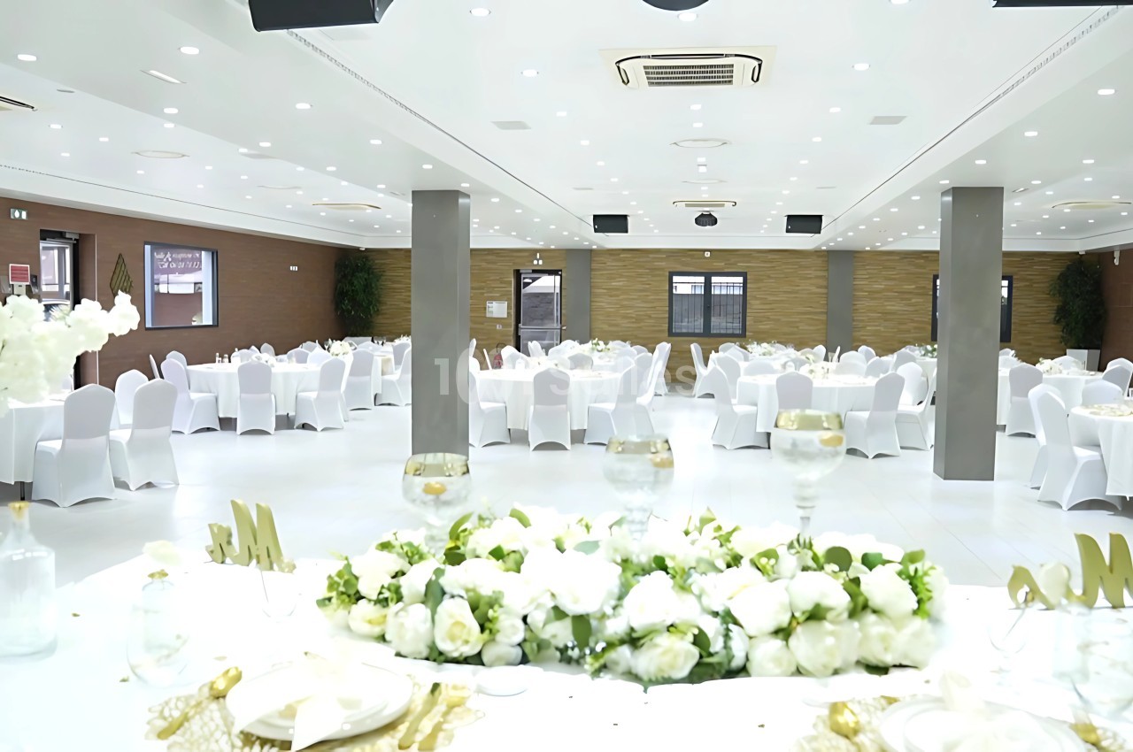 Salle de réception lumineuse avec tables rondes dressées, chaises blanches et décorations florales élégantes.