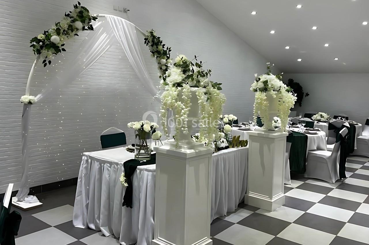 Salle de réception décorée pour un événement, avec arche florale blanche, tables dressées et éclairage tamisé.