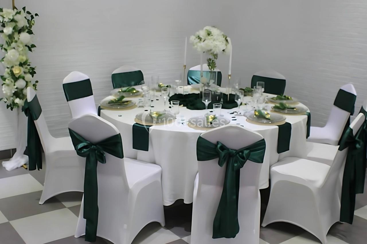 Table ronde décorée pour un événement, avec nappes blanches, chaises ornées de rubans verts et centre de table floral.