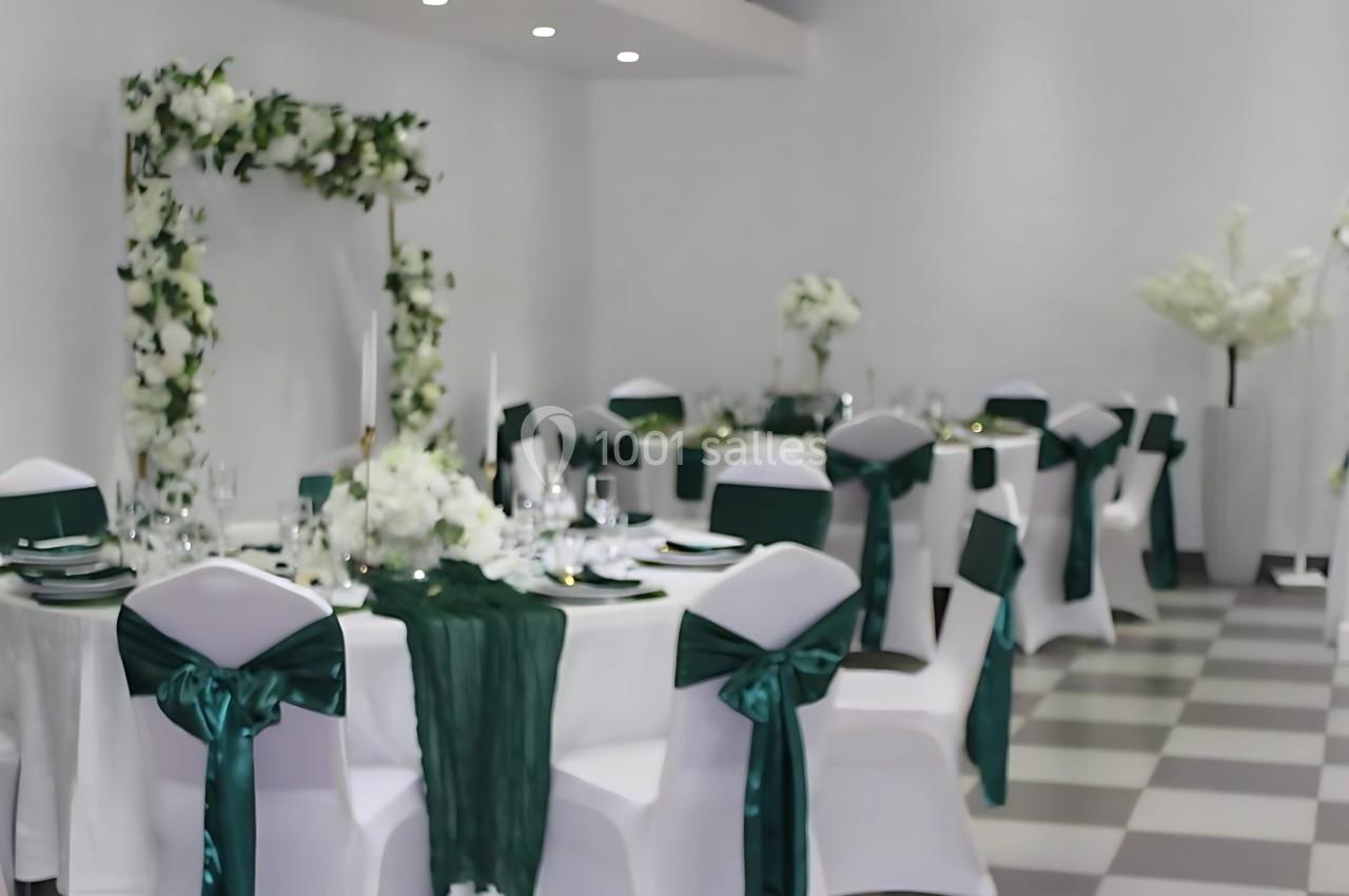 Salle de réception décorée avec des tables rondes, nappes blanches, rubans verts et un arche fleuri en arrière-plan.