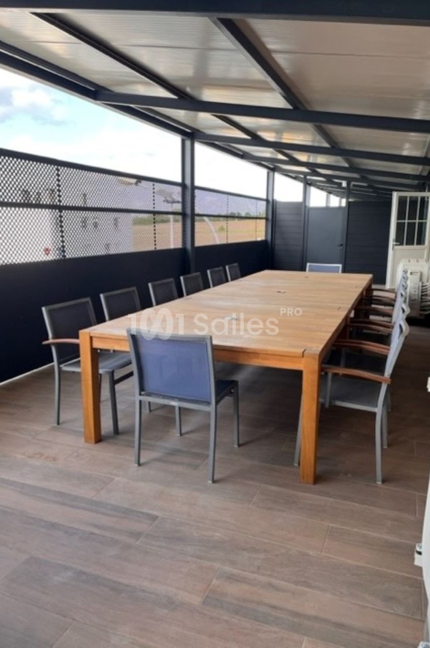 Table en bois rectangulaire entourée de chaises sur une terrasse couverte avec sol en bois et panneaux ajourés.