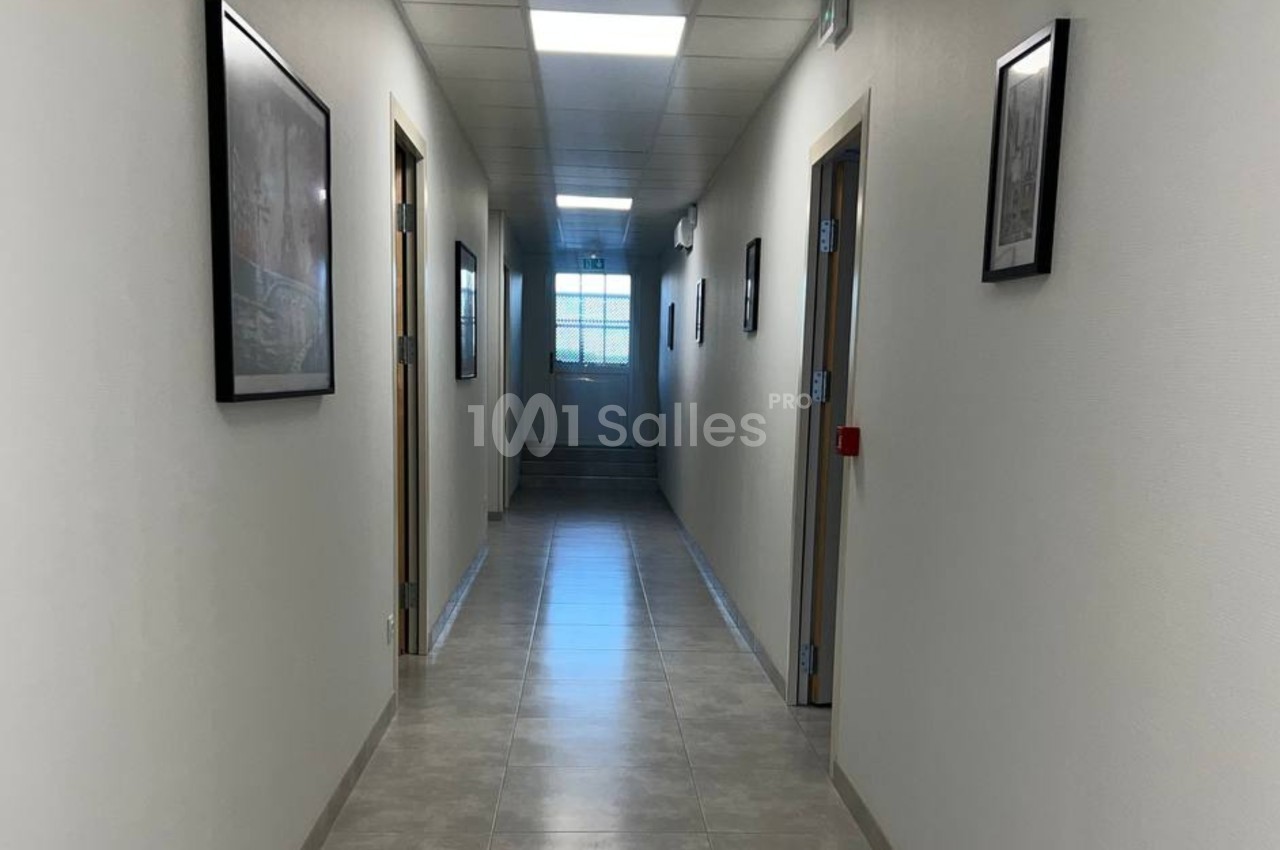 Couloir lumineux avec sol en carrelage, murs blancs ornés de cadres, et une fenêtre au fond.