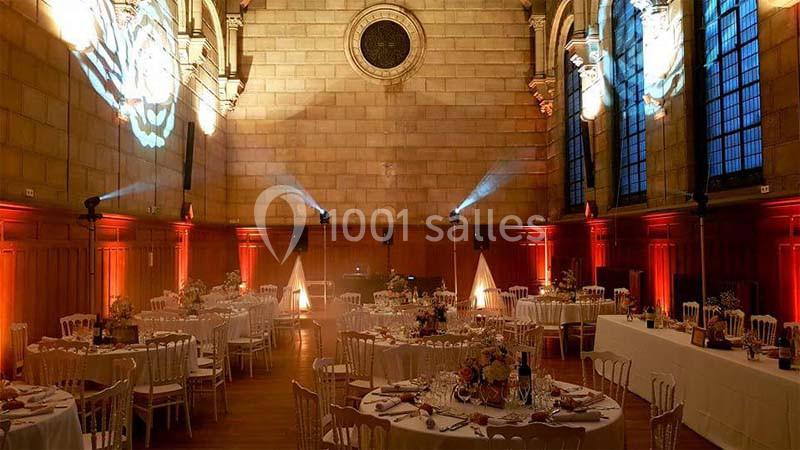 Salle de réception élégante avec tables rondes décorées, éclairage chaleureux et vitraux en arrière-plan.