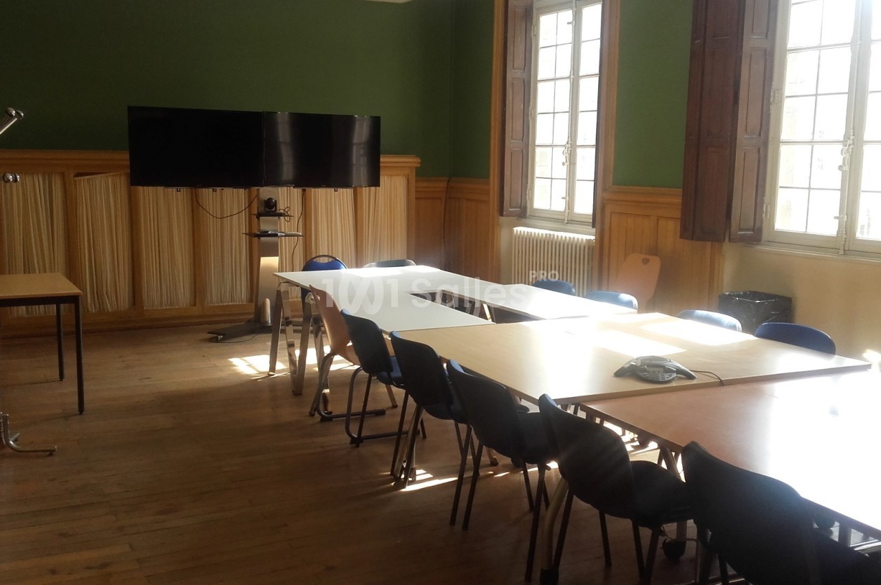 Salle de réunion lumineuse avec tables disposées en rectangle, chaises, écran mural et porte-manteau.