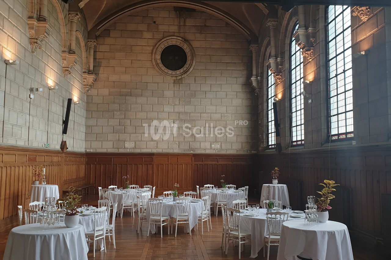 Salle de réception avec des tables rondes dressées, entourées de chaises blanches, dans un décor aux murs en pierre et…