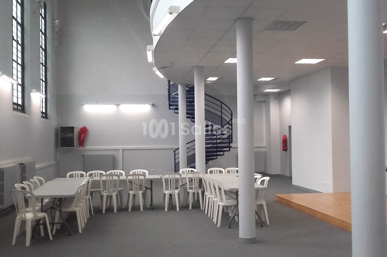 Salle lumineuse avec tables et chaises blanches, escalier en colimaçon bleu et murs clairs.