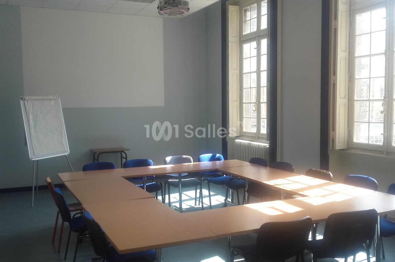 Salle de réunion lumineuse avec tables disposées en U, chaises bleues et un tableau de présentation sur pied.