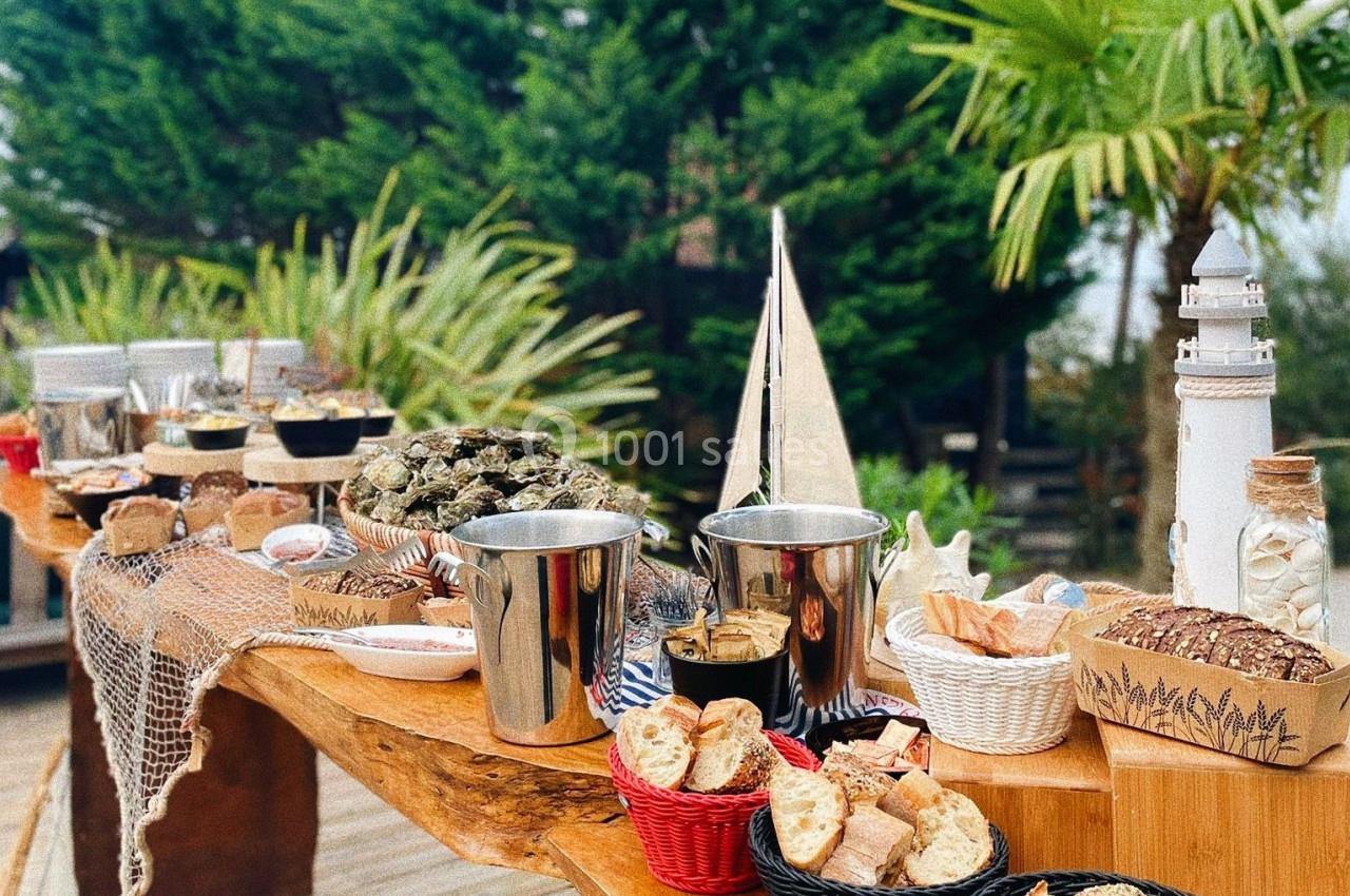 Table en bois garnie de fruits de mer, pains, condiments et décorations marines, entourée de végétation.