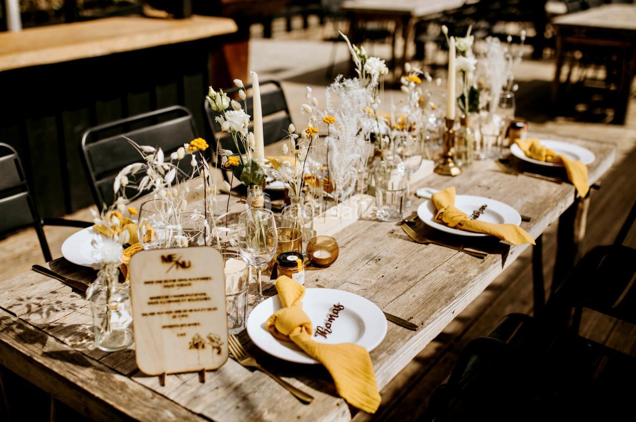 Table en bois décorée pour un repas, avec fleurs, bougies, serviettes jaunes et menus personnalisés.
