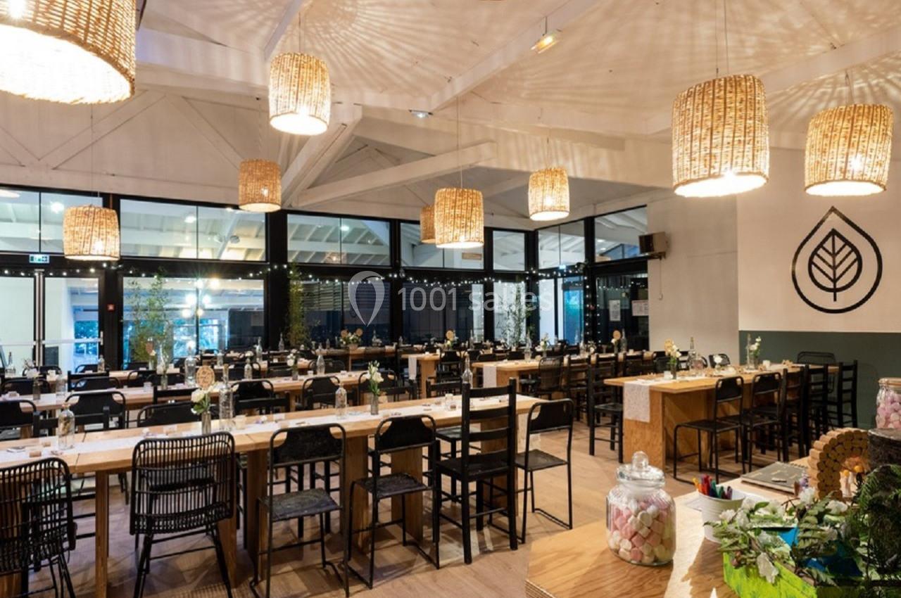 Salle de restaurant lumineuse avec de longues tables en bois, chaises variées et suspensions en osier.