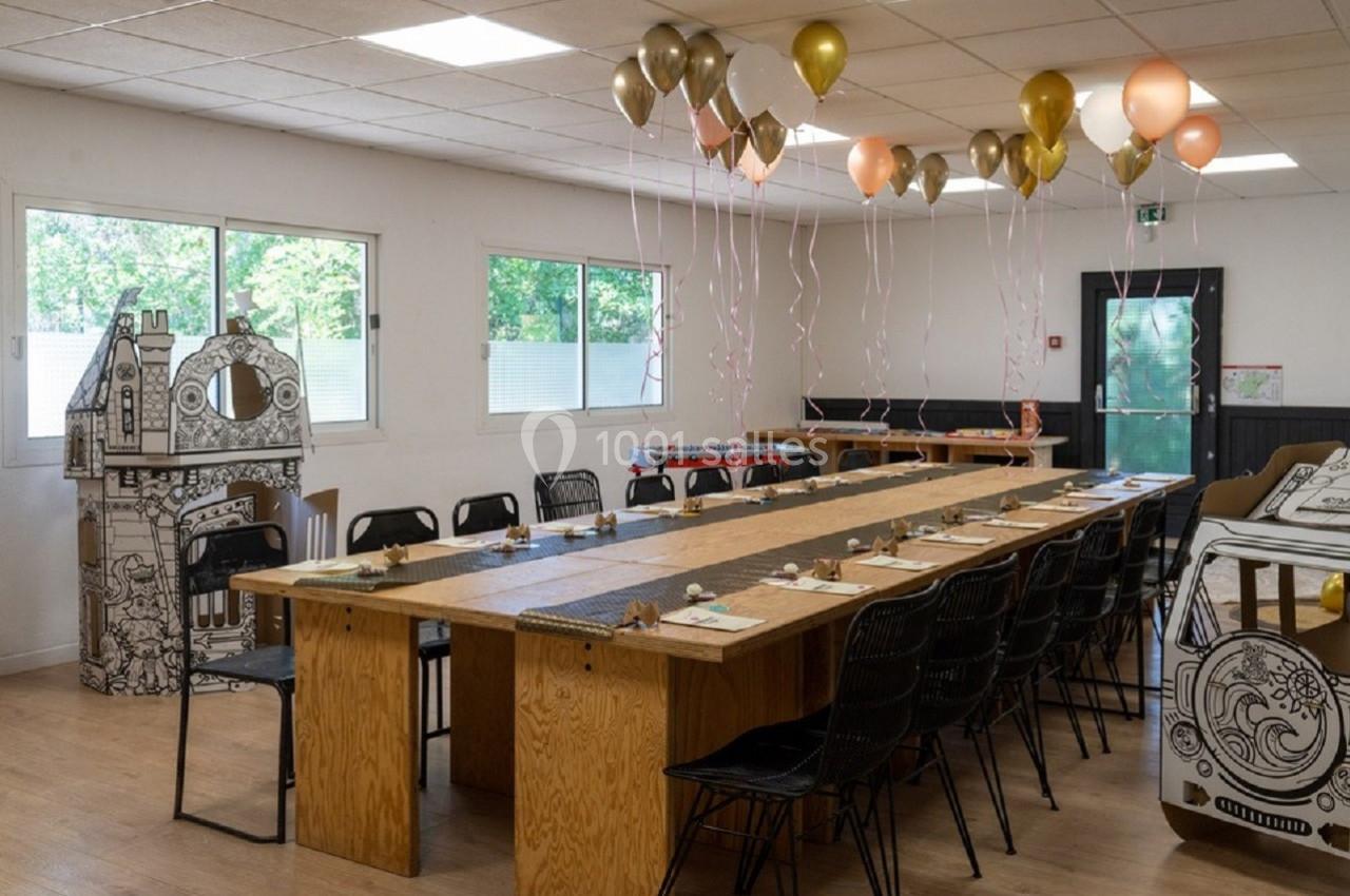 Grande table en bois décorée pour un événement, entourée de chaises, avec des ballons suspendus et des éléments en carton.