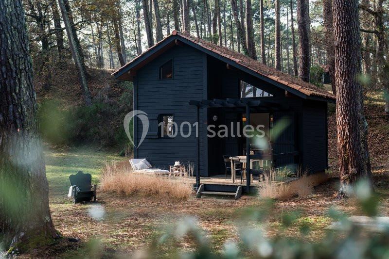 Cabane en bois sombre avec terrasse aménagée, située dans une clairière entourée de pins.