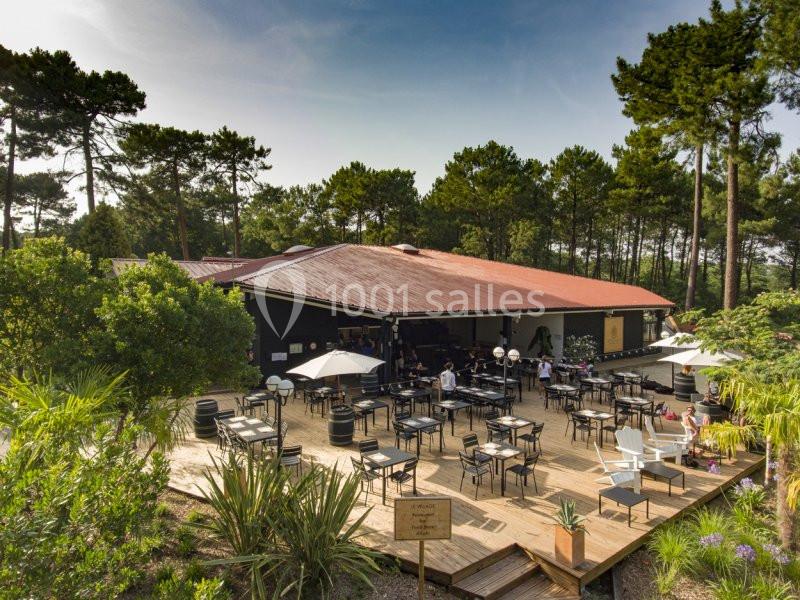 Terrasse en bois d’un restaurant entourée de végétation, avec tables, parasols et chaises sous un ciel dégagé.