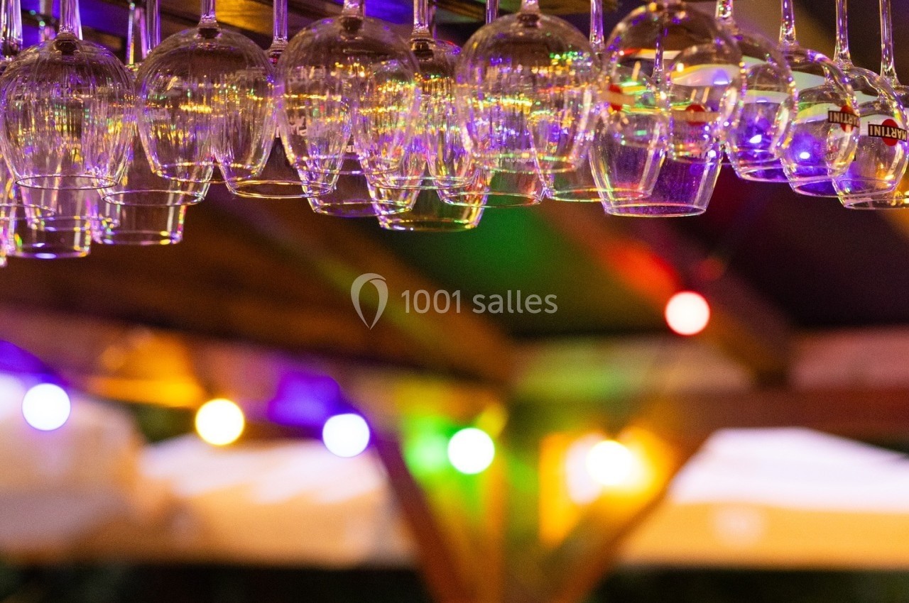 Verres à pied suspendus sous un bar en bois, éclairés par des guirlandes lumineuses colorées en arrière-plan.