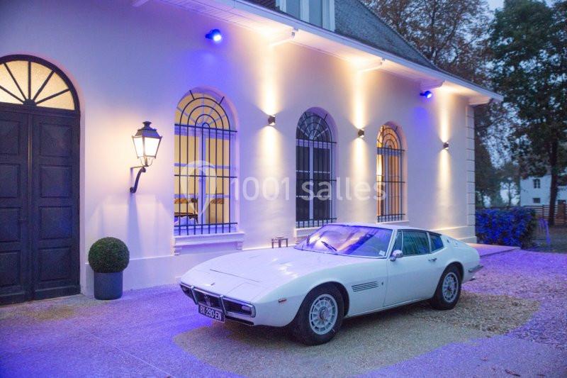 Voiture classique blanche garée devant une maison éclairée avec des fenêtres en arc et des murs clairs.