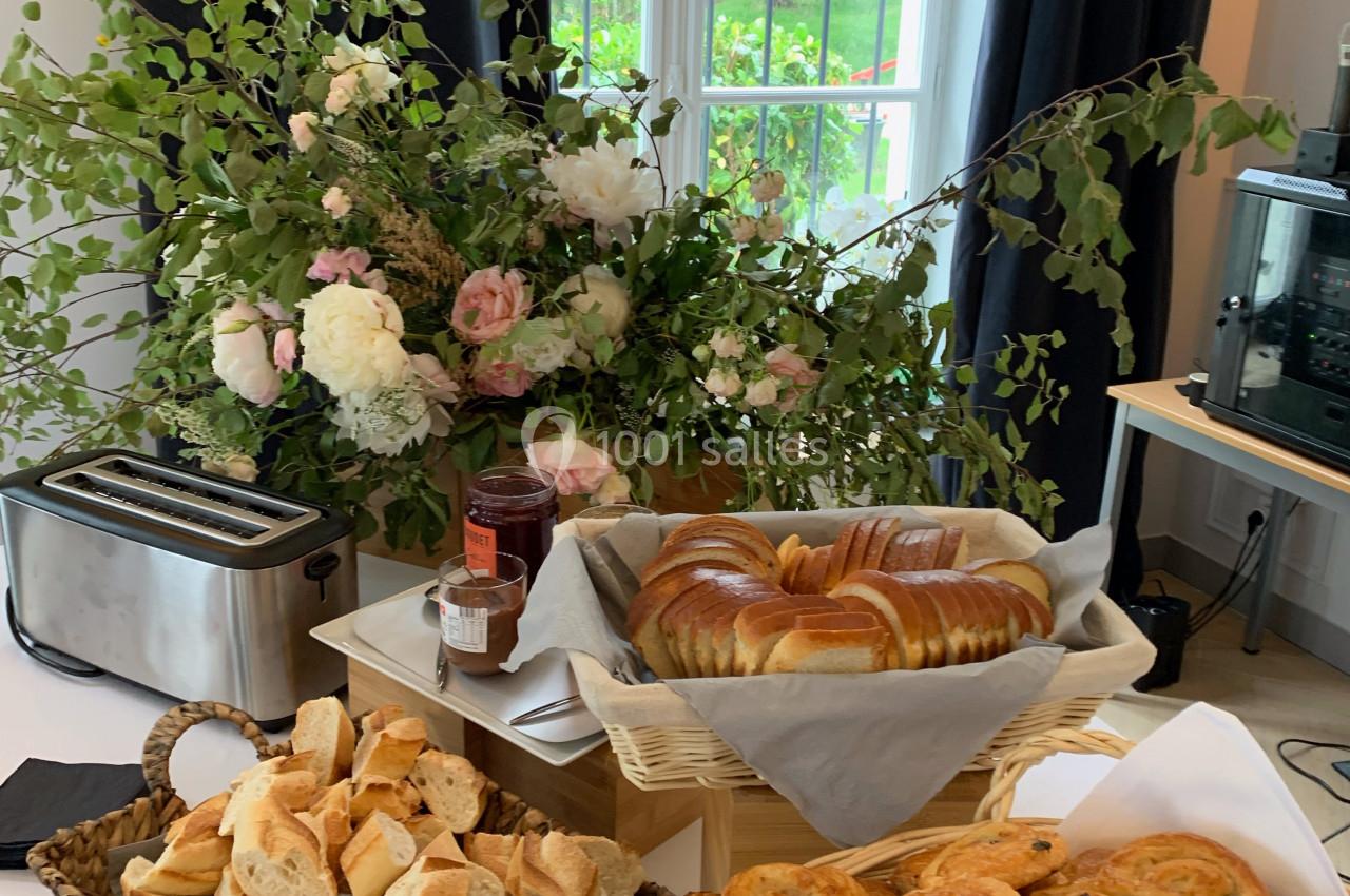 Buffet de petit-déjeuner avec viennoiseries, pain, confiture et grille-pain, devant une fenêtre et des fleurs décoratives.