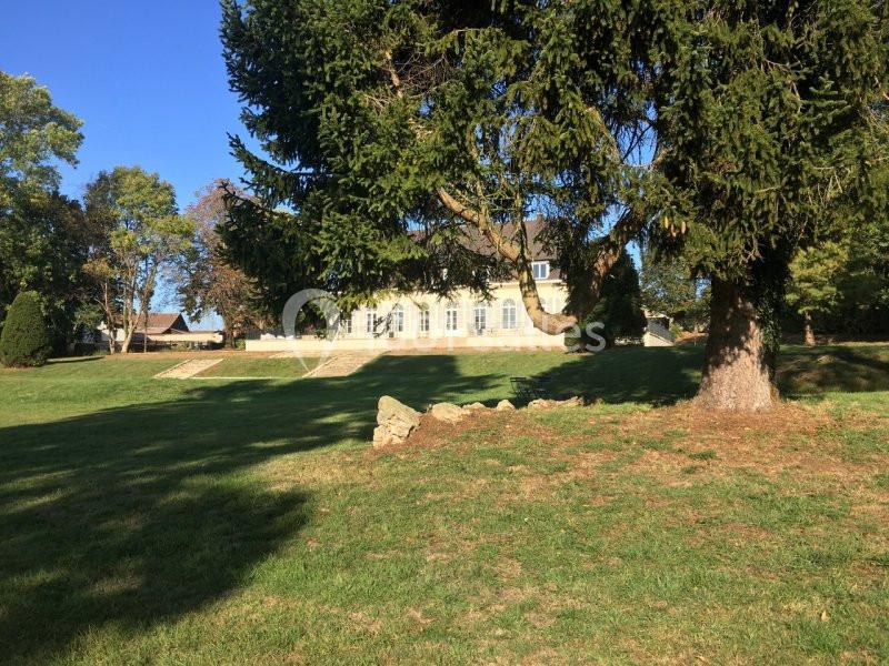 Grande maison blanche entourée d'arbres, vue depuis un jardin avec pelouse et ombre portée au premier plan.
