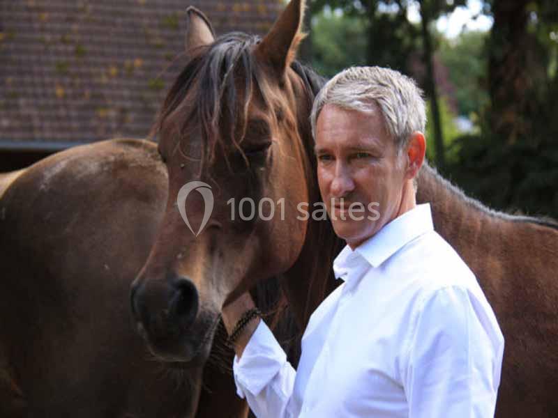 Un homme en chemise blanche pose à côté d'un cheval brun dans un environnement extérieur.