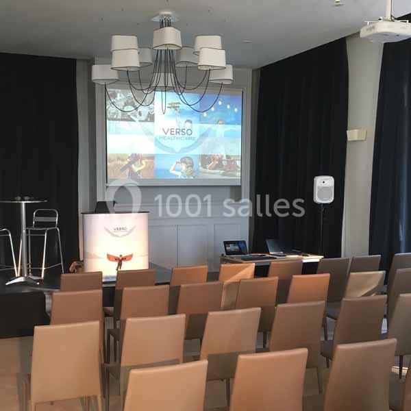 Salle de conférence avec chaises alignées, écran de projection allumé et pupitre en avant-plan.