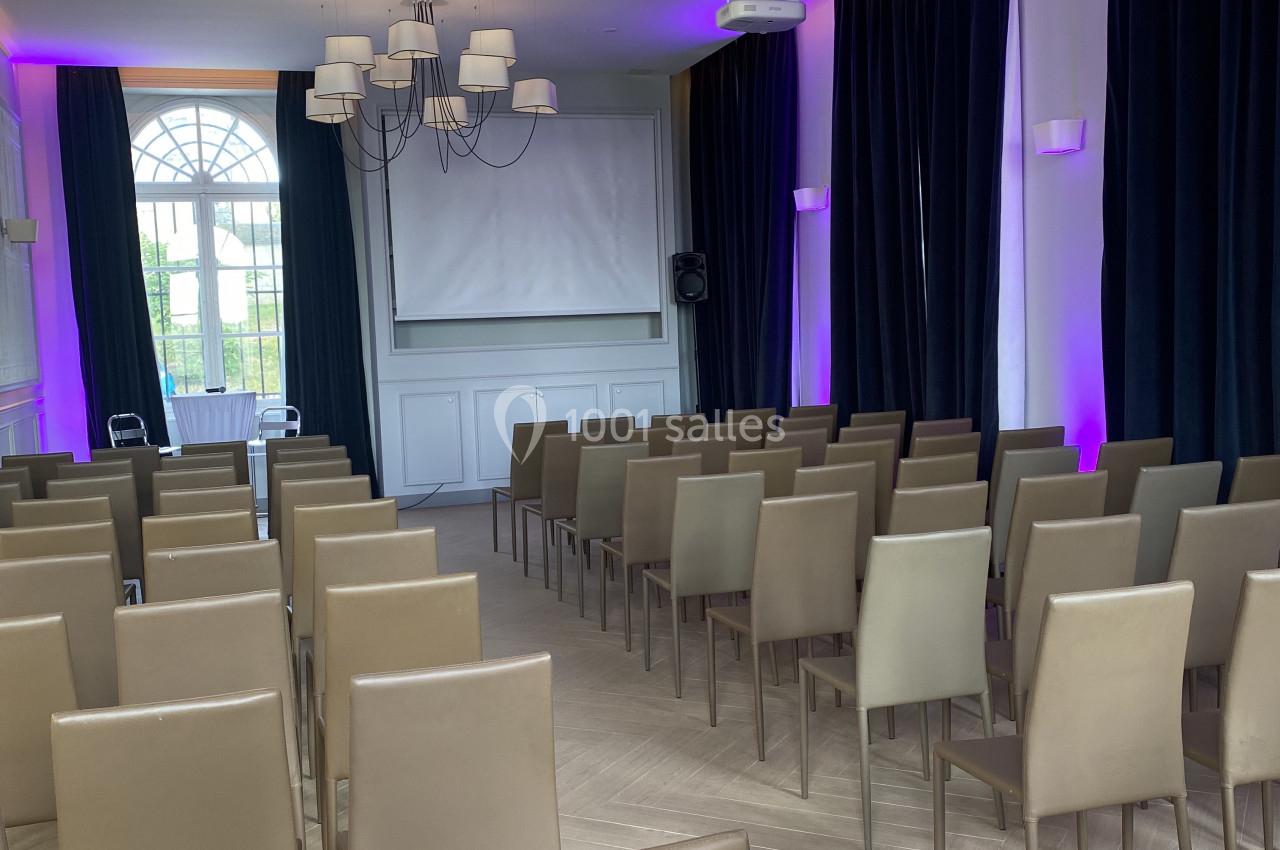 Salle de conférence avec rangées de chaises beige, écran de projection, rideaux sombres et éclairage violet.