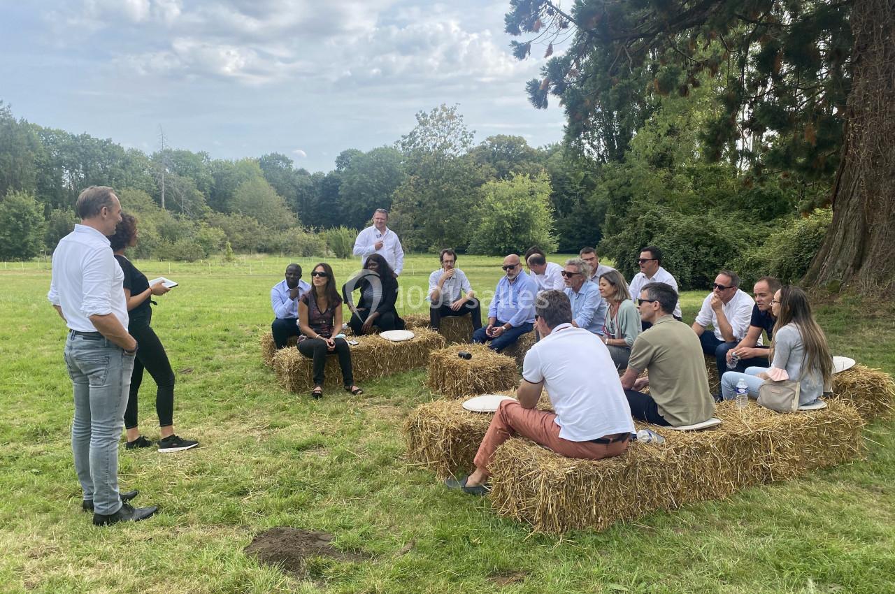 Un groupe de personnes assises sur des bottes de paille en plein air, écoutant deux intervenants debout.