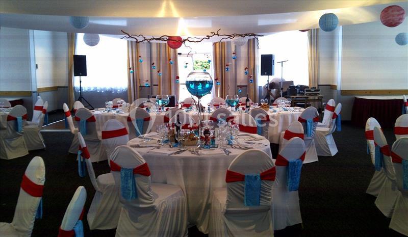 Salle de réception décorée avec des tables rondes, nappes blanches, chaises ornées de rubans rouges et bleus.
