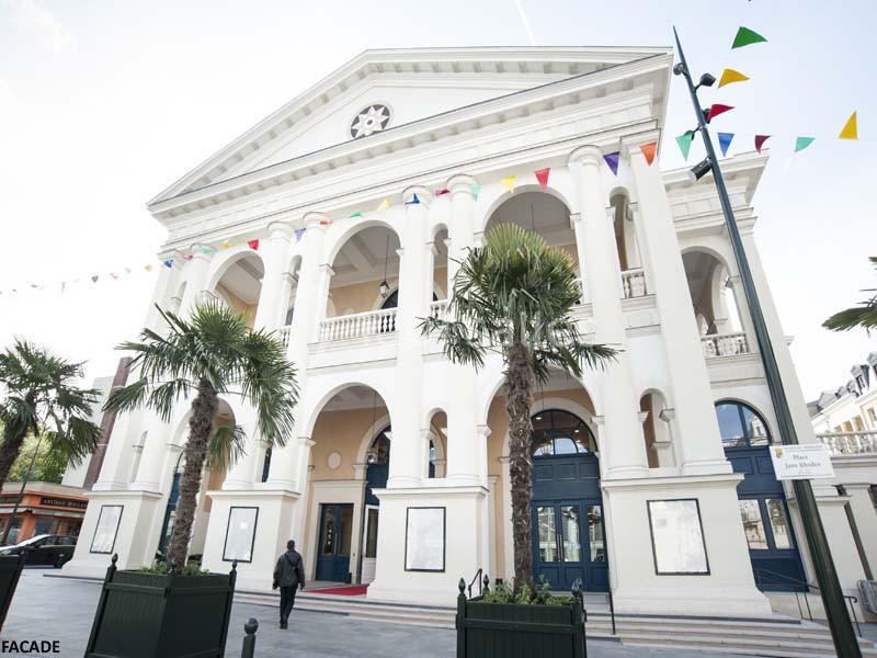 Façade d'un bâtiment néoclassique avec colonnes, décorée de fanions colorés, entourée de palmiers et d'une place pavée.