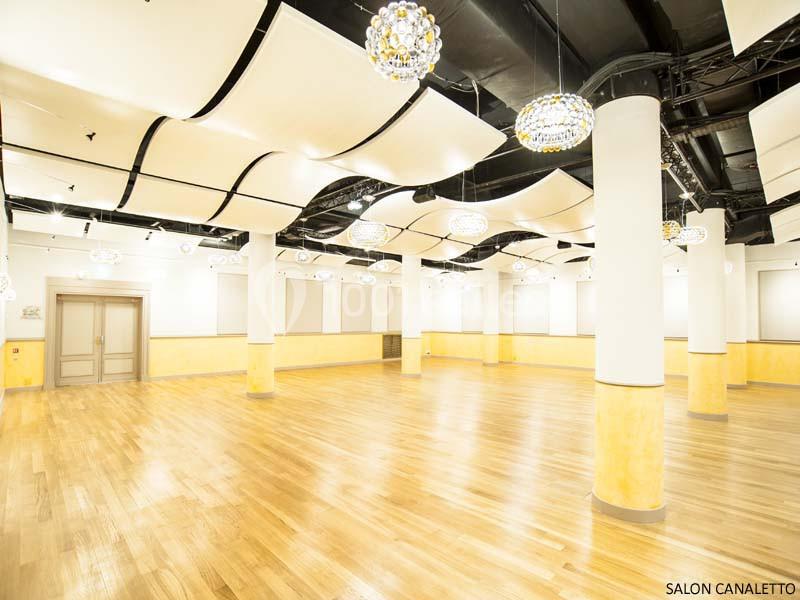 Salle spacieuse avec parquet clair, colonnes blanches, plafonds ondulés et luminaires suspendus.