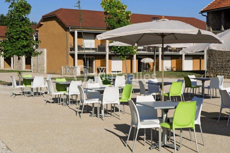 Terrasse extérieure avec tables et chaises blanches et vertes, entourée de bâtiments et d'arbres sous un ciel dégagé.