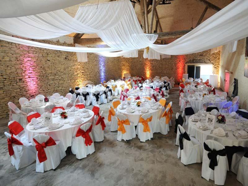 Salle de réception décorée avec des tables rondes, nappes blanches, chaises ornées de nœuds colorés et voilages suspendus.