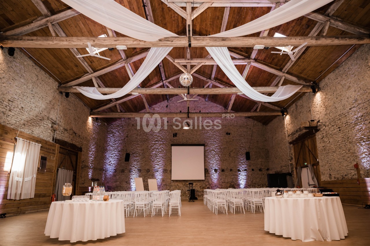 Salle en pierre avec plafond en poutres apparentes, décorée de voilages blancs, tables rondes et chaises blanches.
