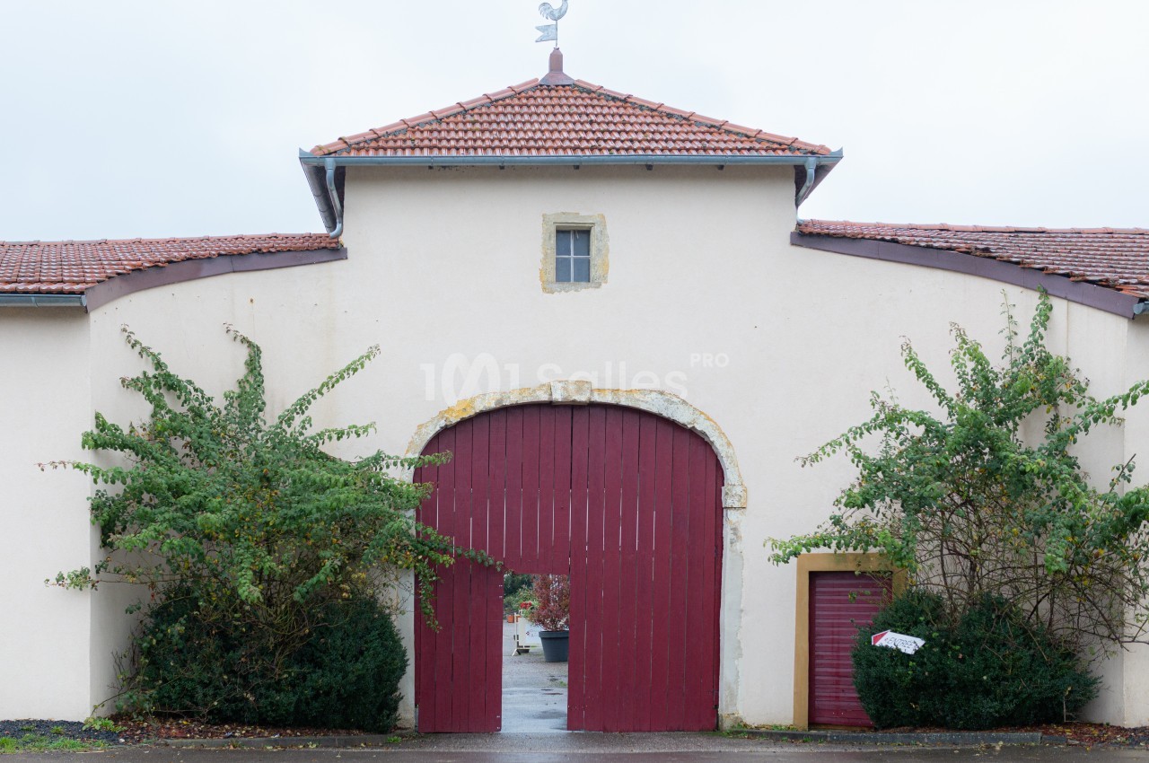 Façade d'un bâtiment ancien avec une grande porte en bois rouge, entourée de végétation et surmontée d'un toit en tuiles.