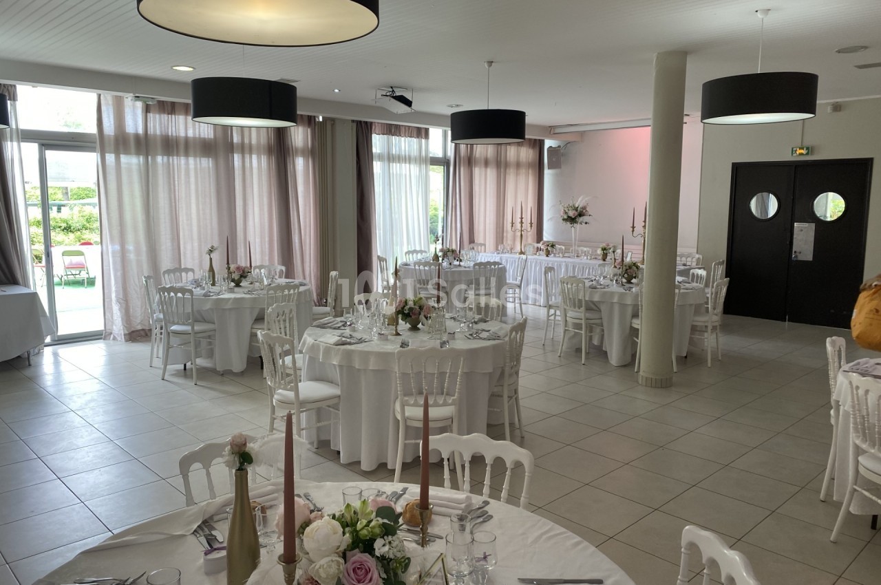 Salle de réception lumineuse avec tables rondes dressées, nappes blanches, décorations florales et grandes baies vitrées.
