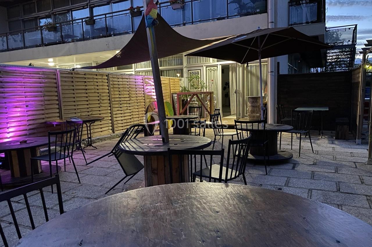 Terrasse extérieure avec tables en bois, chaises noires et éclairage d'ambiance violet en arrière-plan.