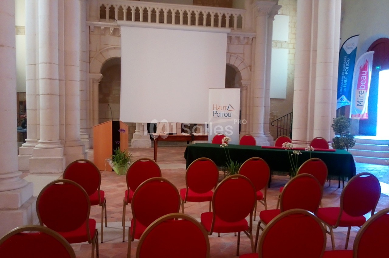 Salle avec des chaises rouges alignées, une table avec des micros, un écran de projection et des bannières promotionnelles.