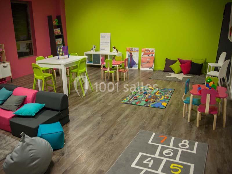 Salle de jeux colorée avec tables, chaises, tapis de jeu, coussins et mobilier adapté aux enfants.