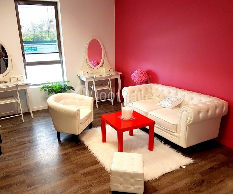 Salon lumineux avec canapé et fauteuil blancs, table rouge, tapis blanc, murs rouges et coiffeuses près d'une fenêtre.