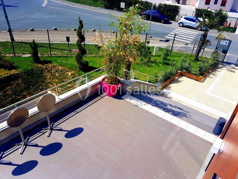 Terrasse ensoleillée avec carrelage, deux tables rondes pliées, une grande plante en pot rose et vue sur une rue.