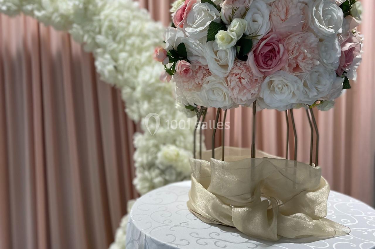 Arrangement floral rond avec des roses blanches et roses sur un support doré, devant un rideau rose et une arche fleurie…