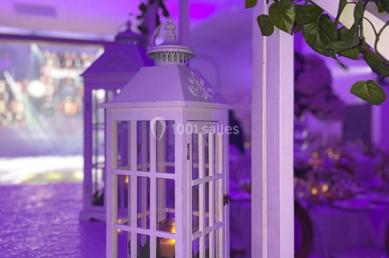 Lanterne blanche décorative avec une bougie allumée, entourée de feuillage, dans une salle éclairée par des lumières…