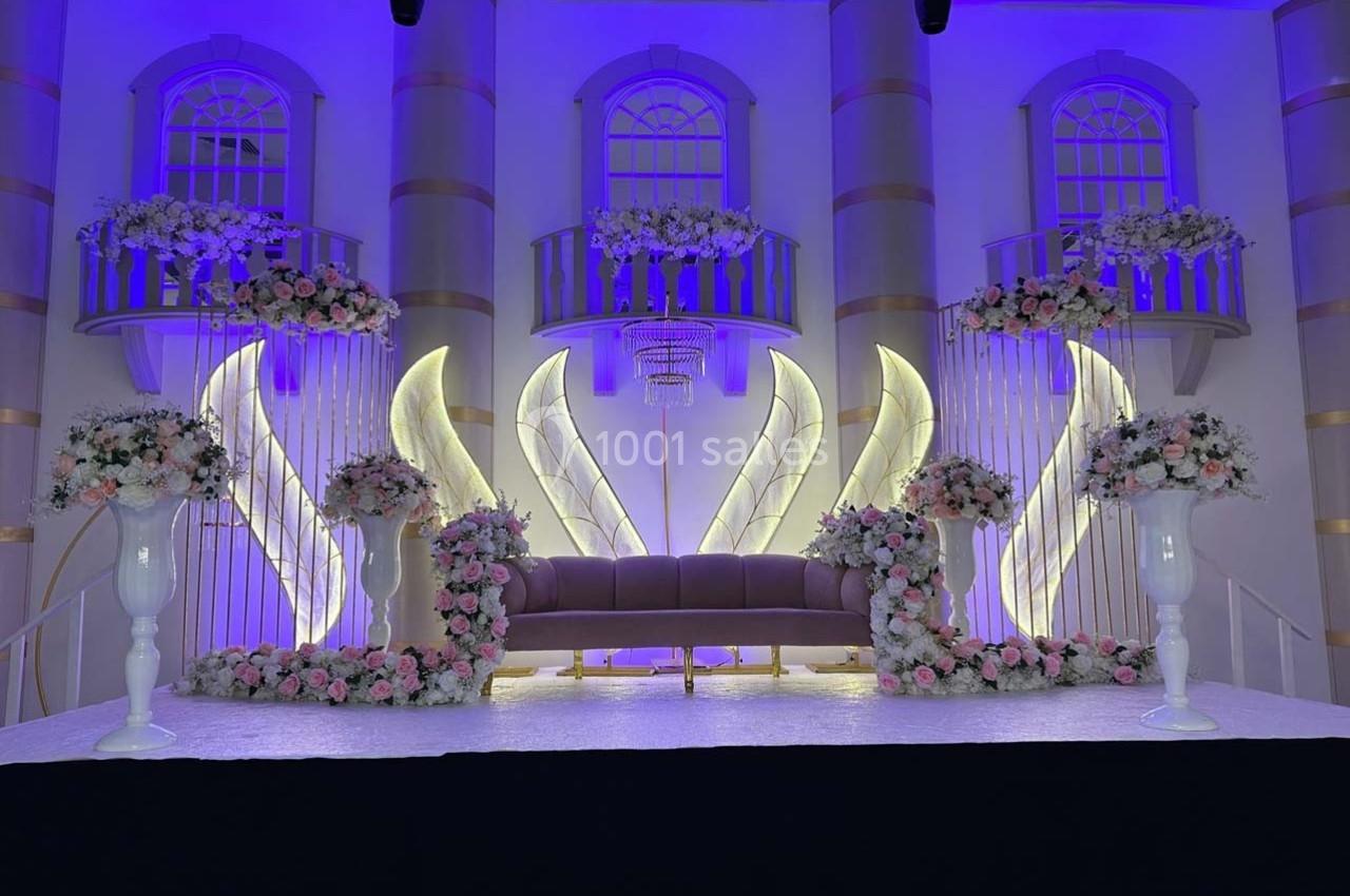 Décor de scène élégante avec canapé central, fleurs blanches et roses, éclairage violet et arches lumineuses.