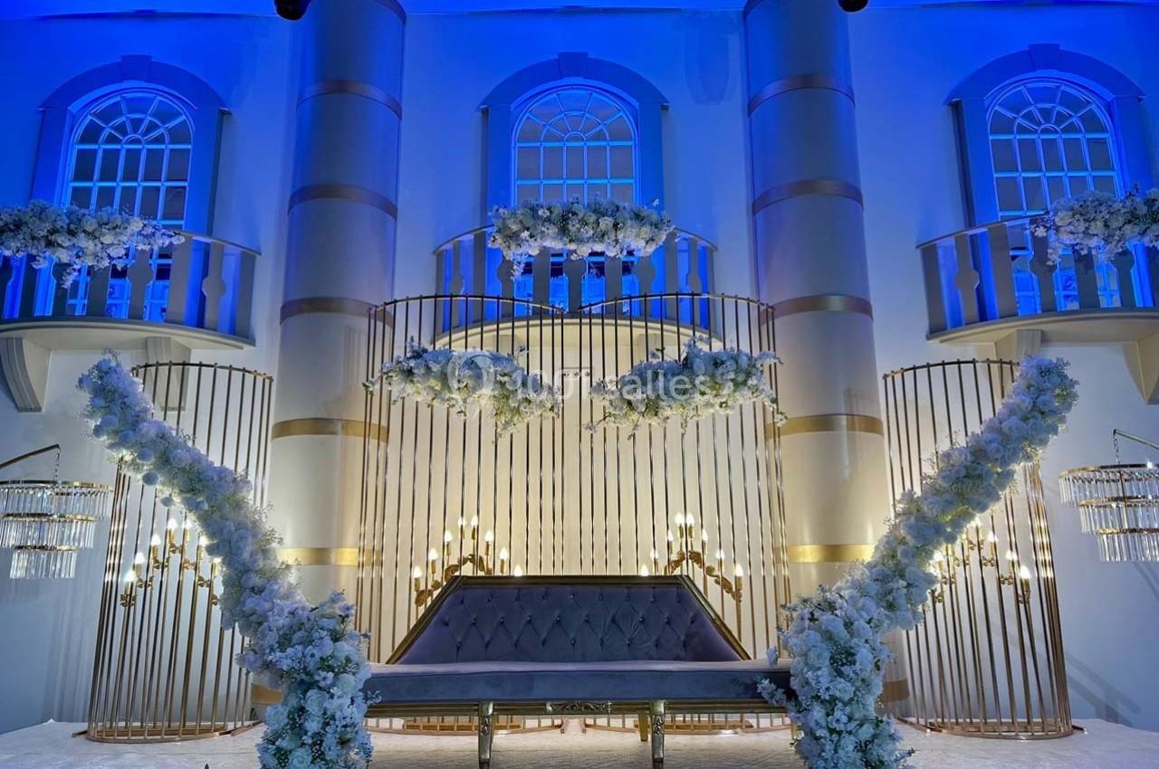 Décor de mariage avec arche dorée, fleurs blanches et éclairage bleu dans une salle élégante.