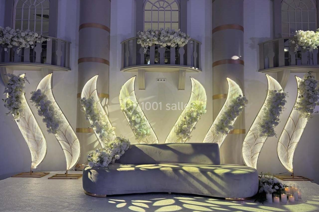 Décor élégant avec un canapé gris, des fleurs blanches et des structures lumineuses en forme de feuilles devant une façade.