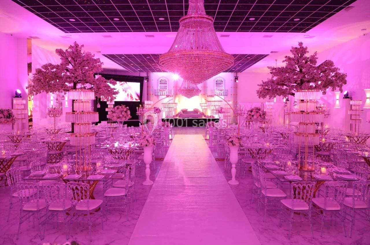 Salle de réception décorée avec des tons roses, des tables élégantes, des chandeliers et un grand lustre central.