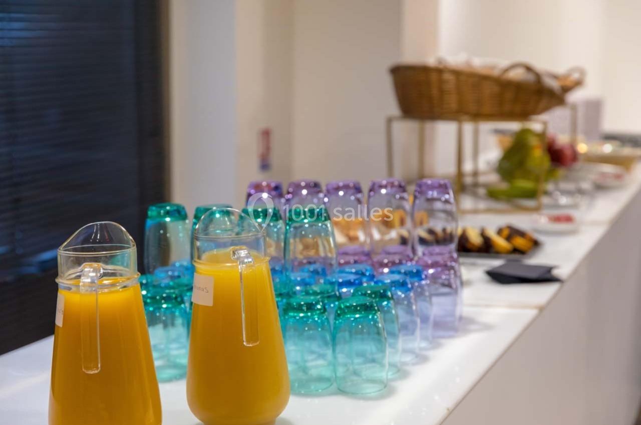 Carafes de jus d'orange et verres colorés alignés sur une table, avec un panier de viennoiseries en arrière-plan.