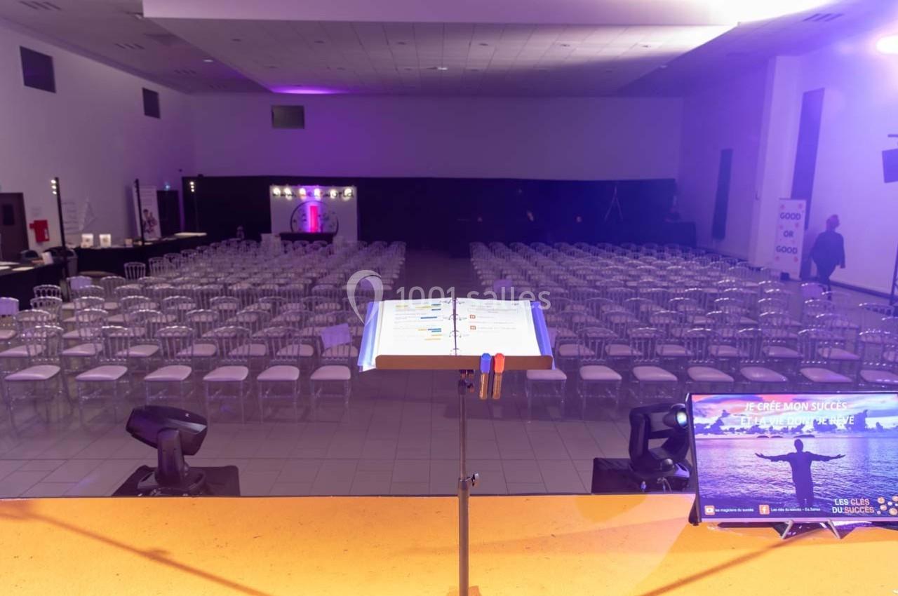 Salle de conférence vide avec des rangées de chaises alignées, vue depuis un pupitre équipé d'un écran et de documents.