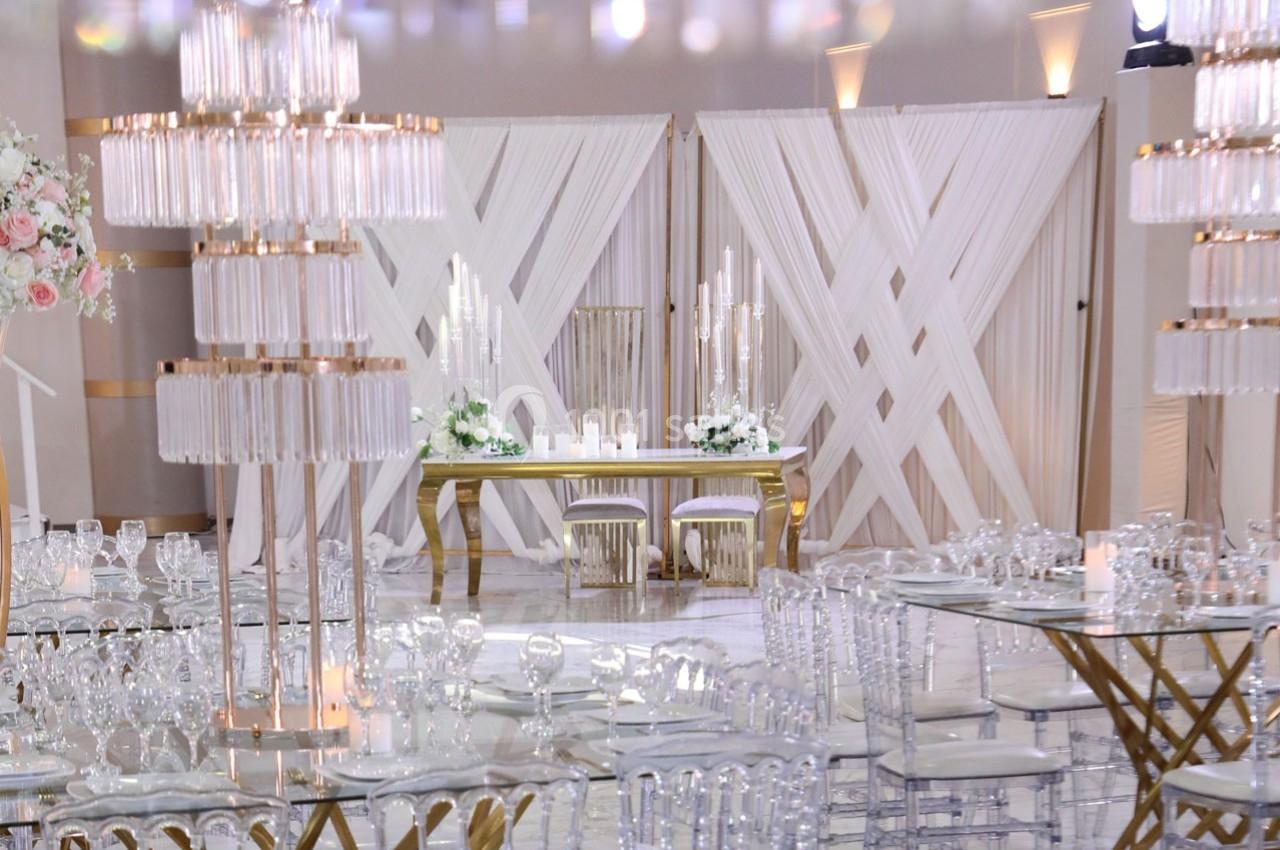 Salle de réception élégante avec table d'honneur décorée de bougies, fleurs blanches et chaises transparentes.