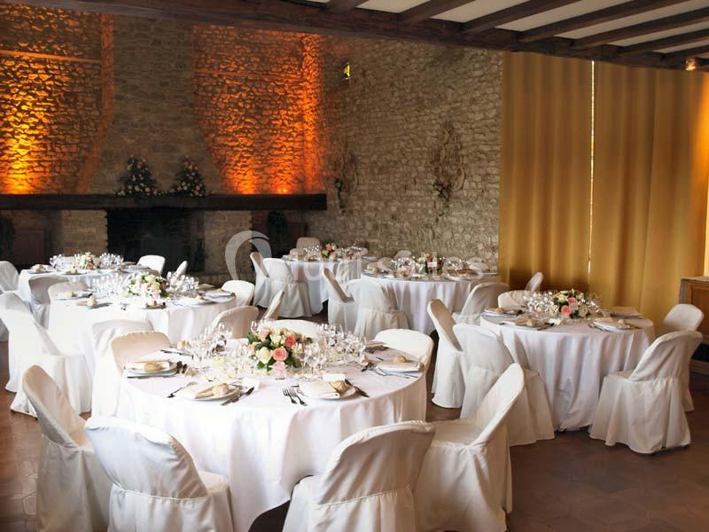 Salle de réception avec des tables rondes dressées, nappes blanches et décorations florales, éclairage chaleureux.