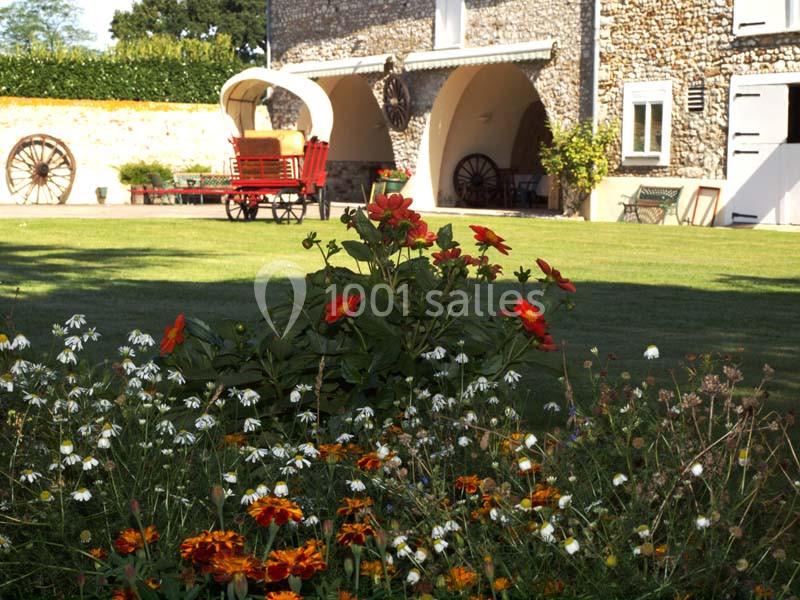 Jardin fleuri avec des marguerites et des dahlias rouges, devant une maison en pierre avec une charrette rouge.